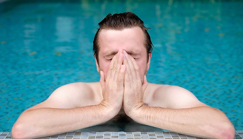 Hombre en una piscina tapándose los ojos con irritación ocular tras el contacto con cloro.webp